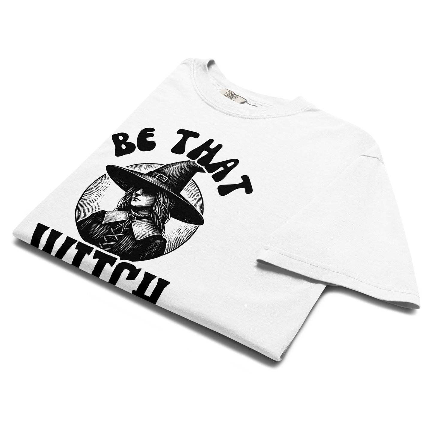 Folded white t-shirt with a vintage witch illustration wearing a wide hat and the phrase ‘Be That Witch’ printed in bold black letters.
