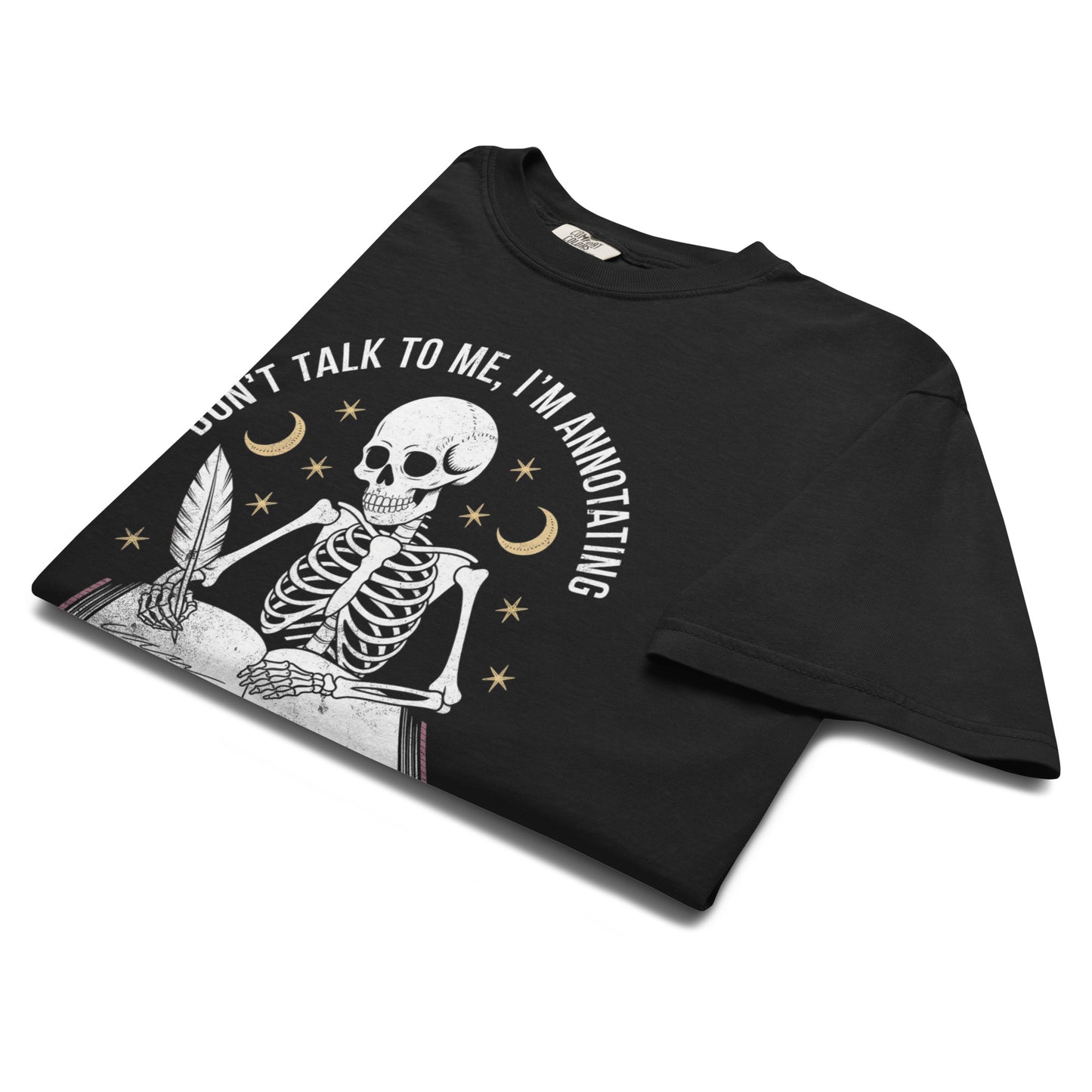 “Folded black t-shirt featuring a skeleton writing in a book under crescent moons and stars with text ‘Don’t talk to me, I’m annotating.