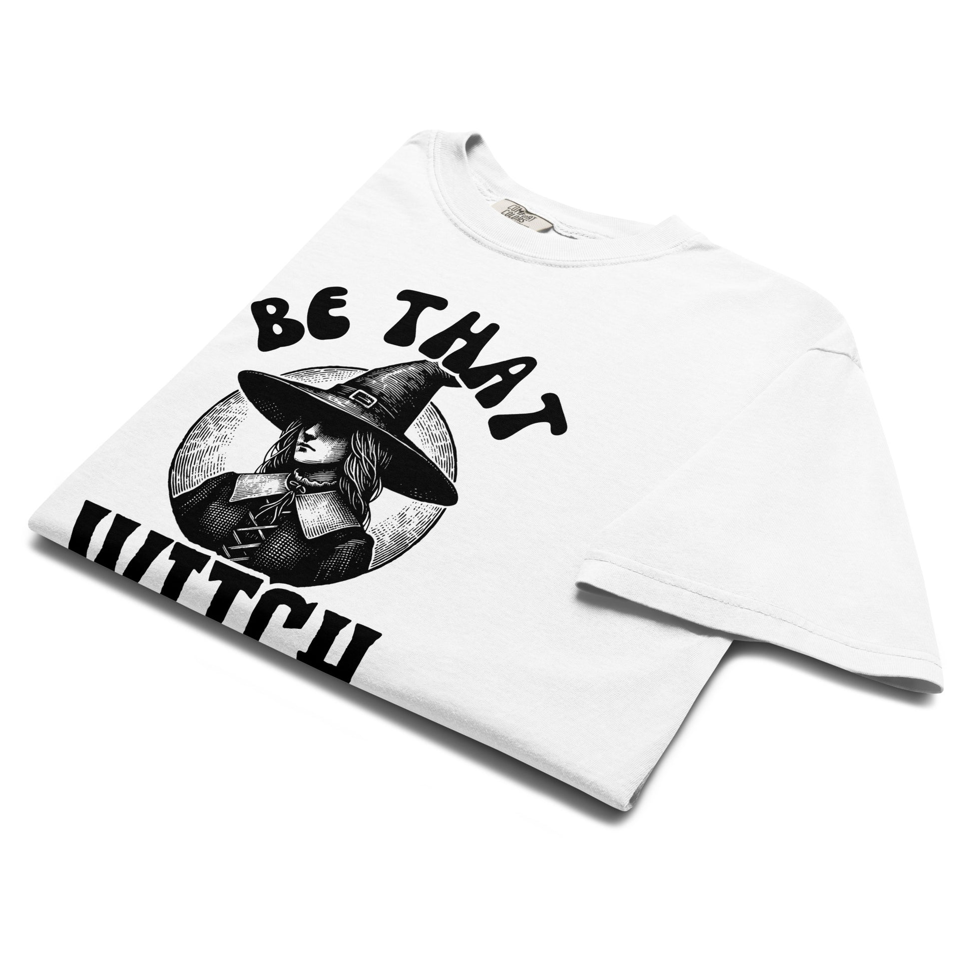 Folded white t-shirt with a vintage witch illustration wearing a wide hat and the phrase ‘Be That Witch’ printed in bold black letters.
