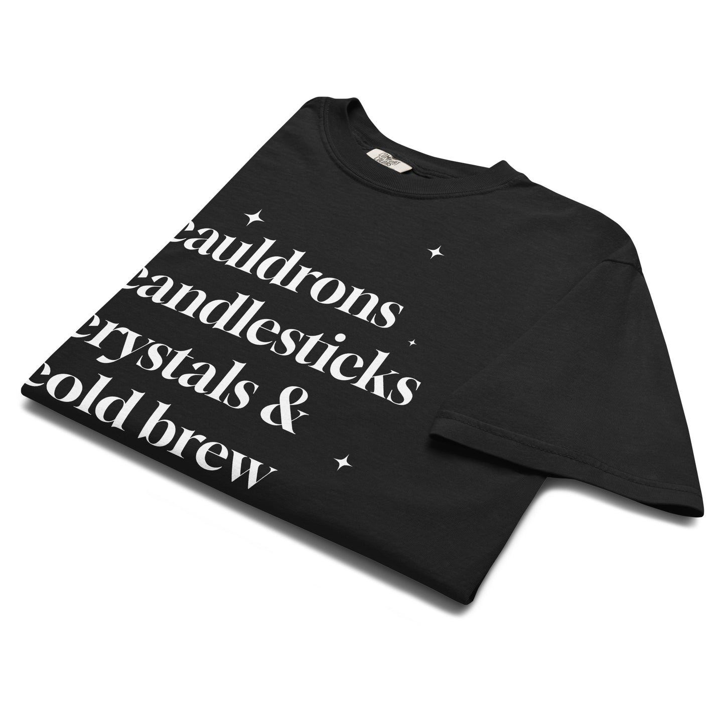 Folded black t-shirt showing white text that reads ‘cauldrons, candlesticks, crystals & cold brew’ in classic serif font with small stars.