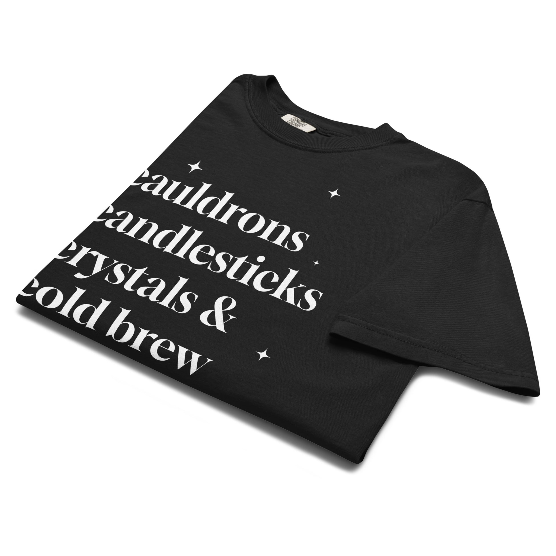 Folded black t-shirt showing white text that reads ‘cauldrons, candlesticks, crystals & cold brew’ in classic serif font with small stars.