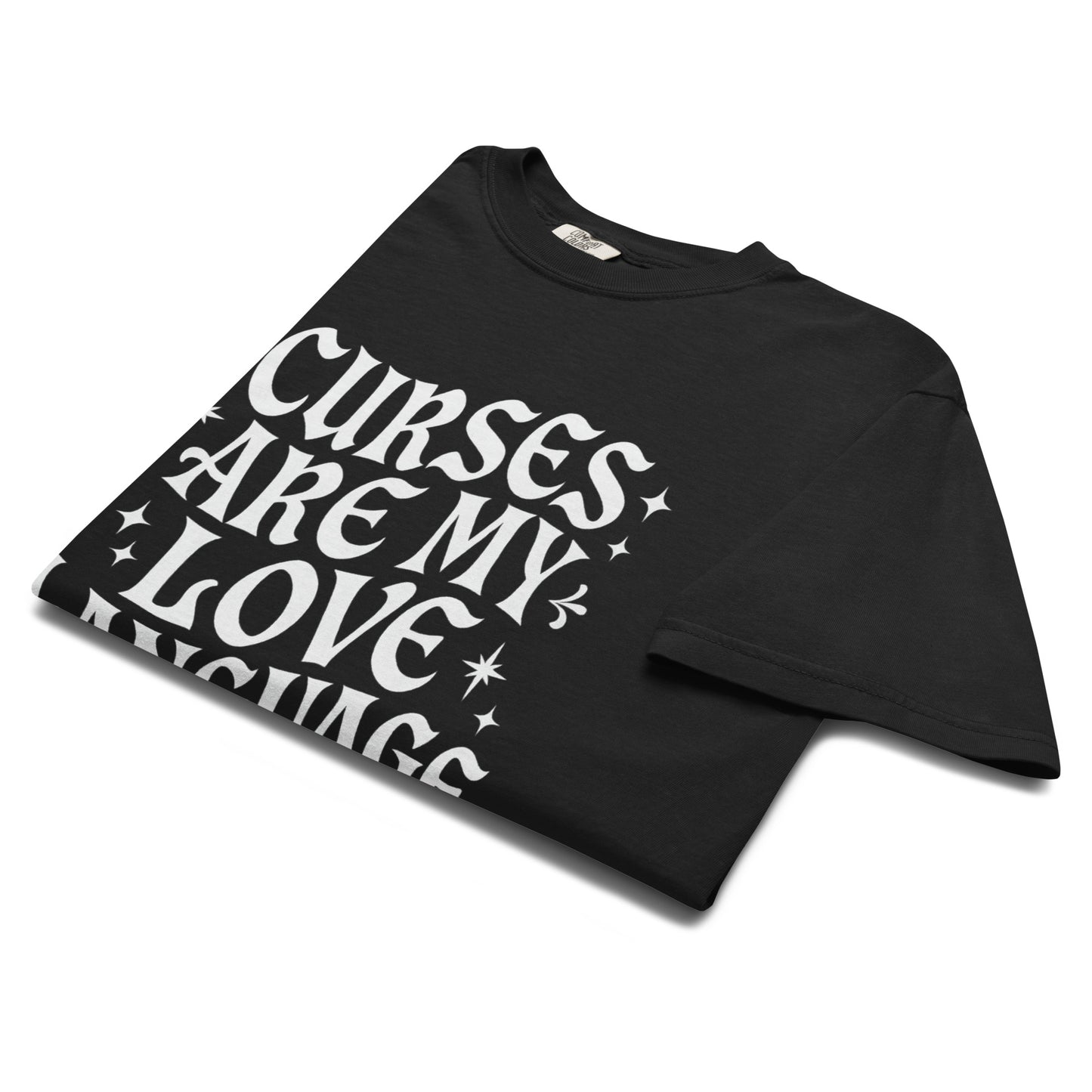 Folded black t-shirt showing bold white gothic text reading ‘Curses Are My Love Language’ with scattered star accents.