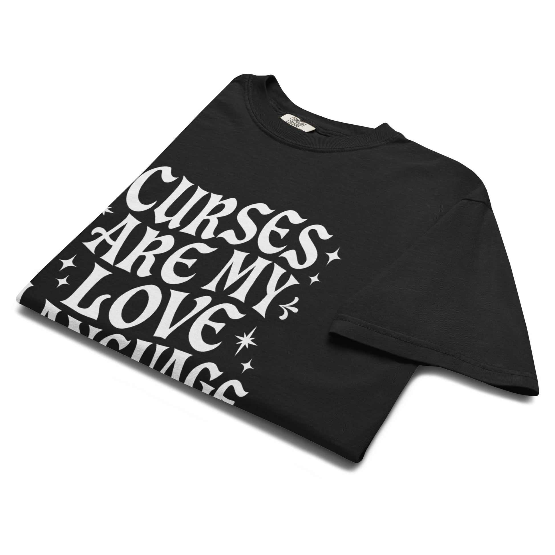 Folded black t-shirt showing bold white gothic text reading ‘Curses Are My Love Language’ with scattered star accents.