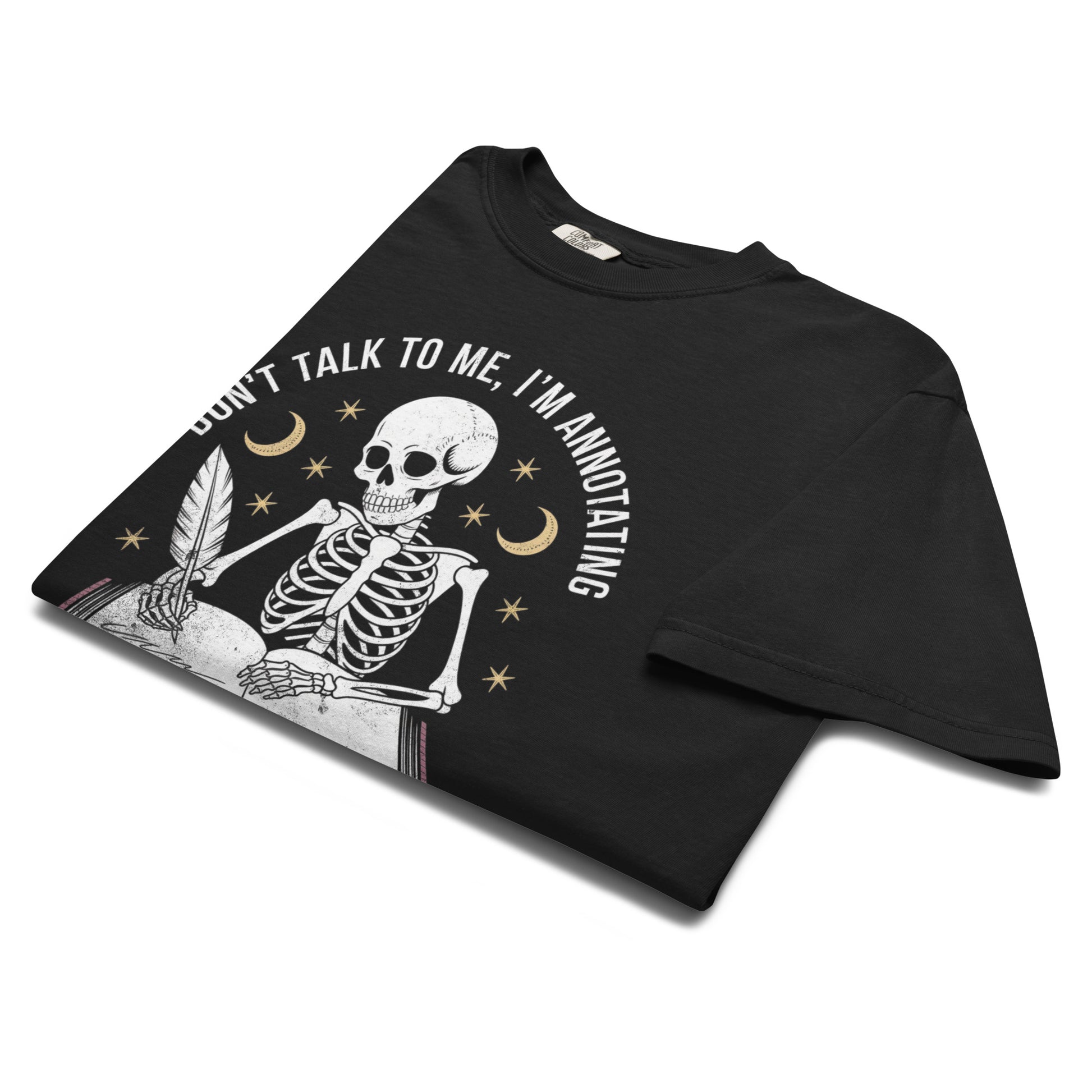 “Folded black t-shirt featuring a skeleton writing in a book under crescent moons and stars with text ‘Don’t talk to me, I’m annotating.