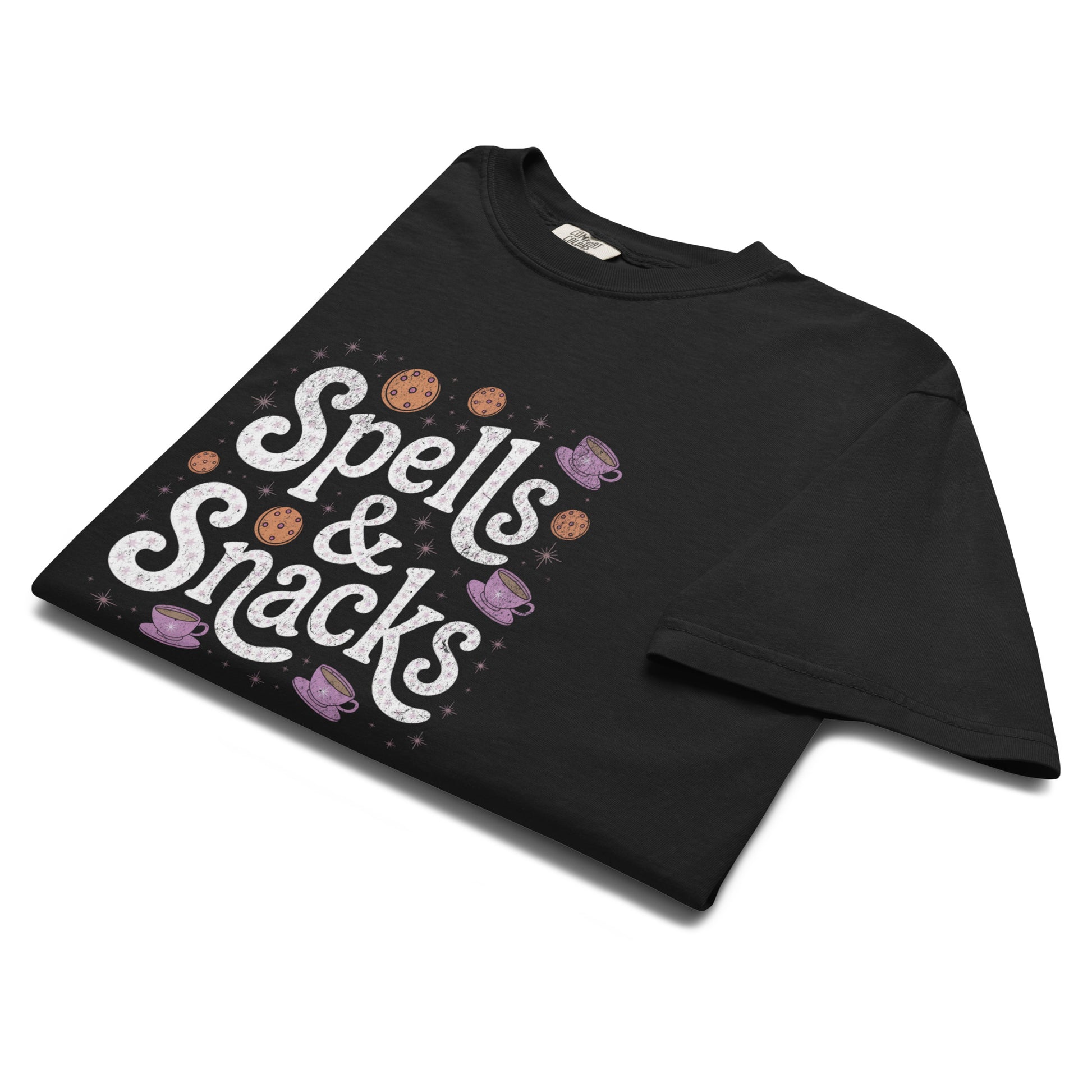 Folded black “Spells & Snacks” t-shirt flat lay showing playful witchy lettering and illustrated treats, perfect for soft goths and magical food lovers alike.