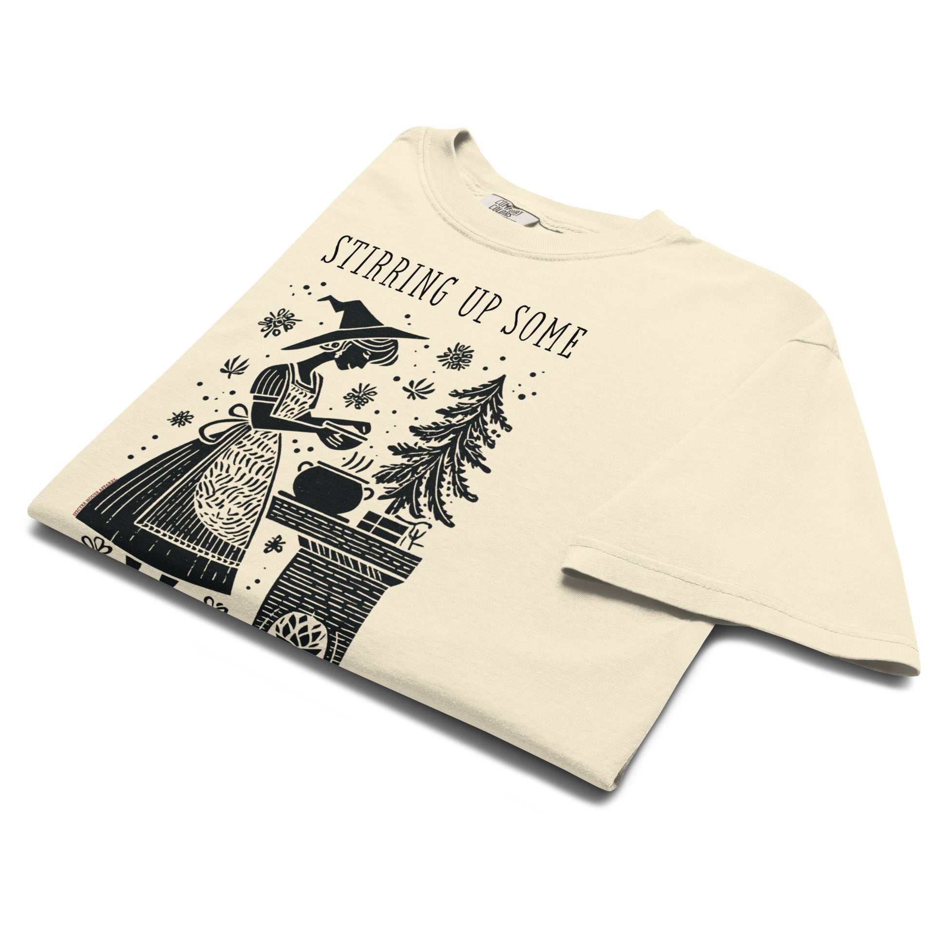 Folded cream “Stirring Up Some Yule Magic” t-shirt flat lay showing vintage witch and cauldron design, ideal for cozy goths and Yule season celebrations.