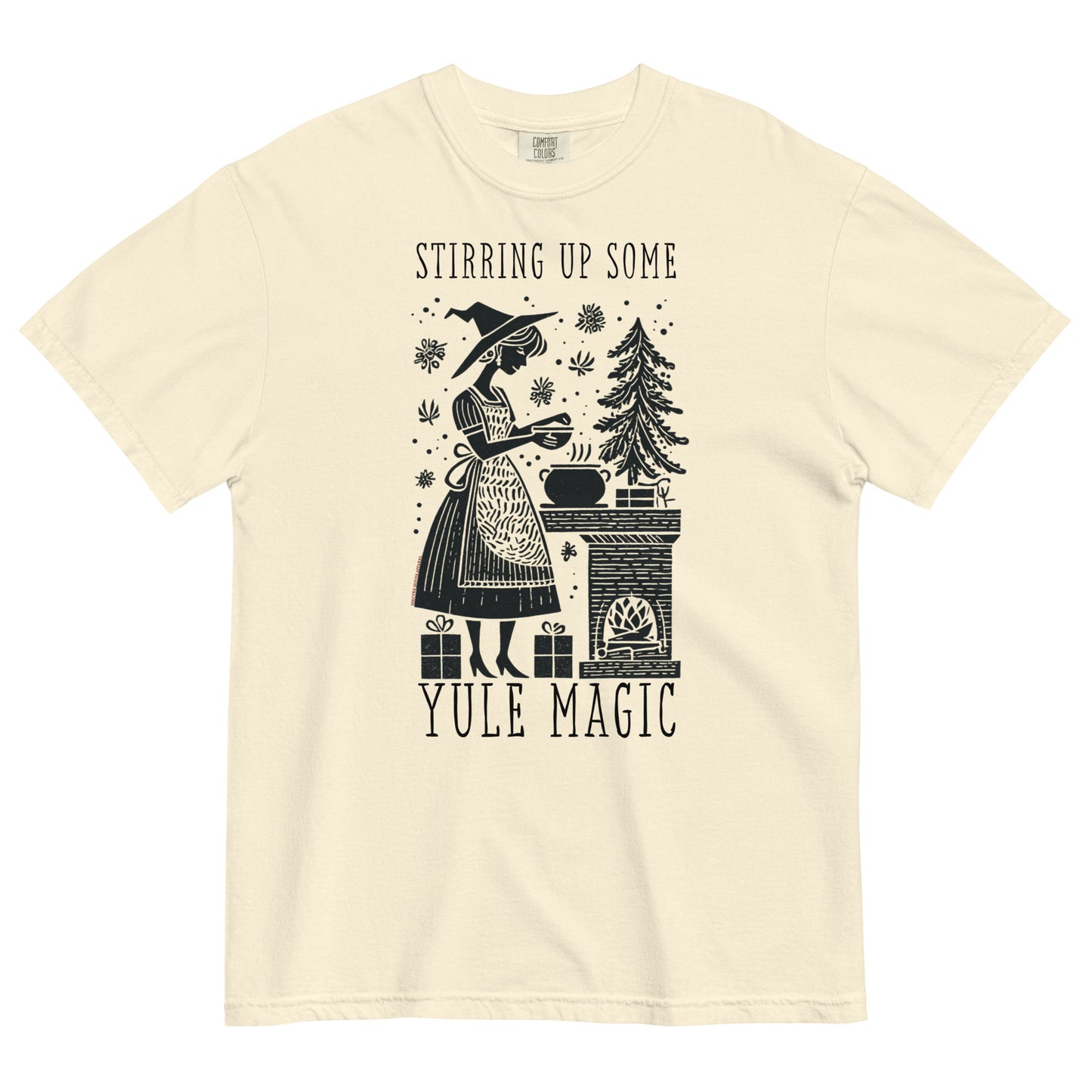Cream t-shirt lay flat featuring vintage illustration of a witch stirring a cauldron beside a pine tree with text “Stirring Up Some Yule Magic,” a cozy witchy holiday tee.