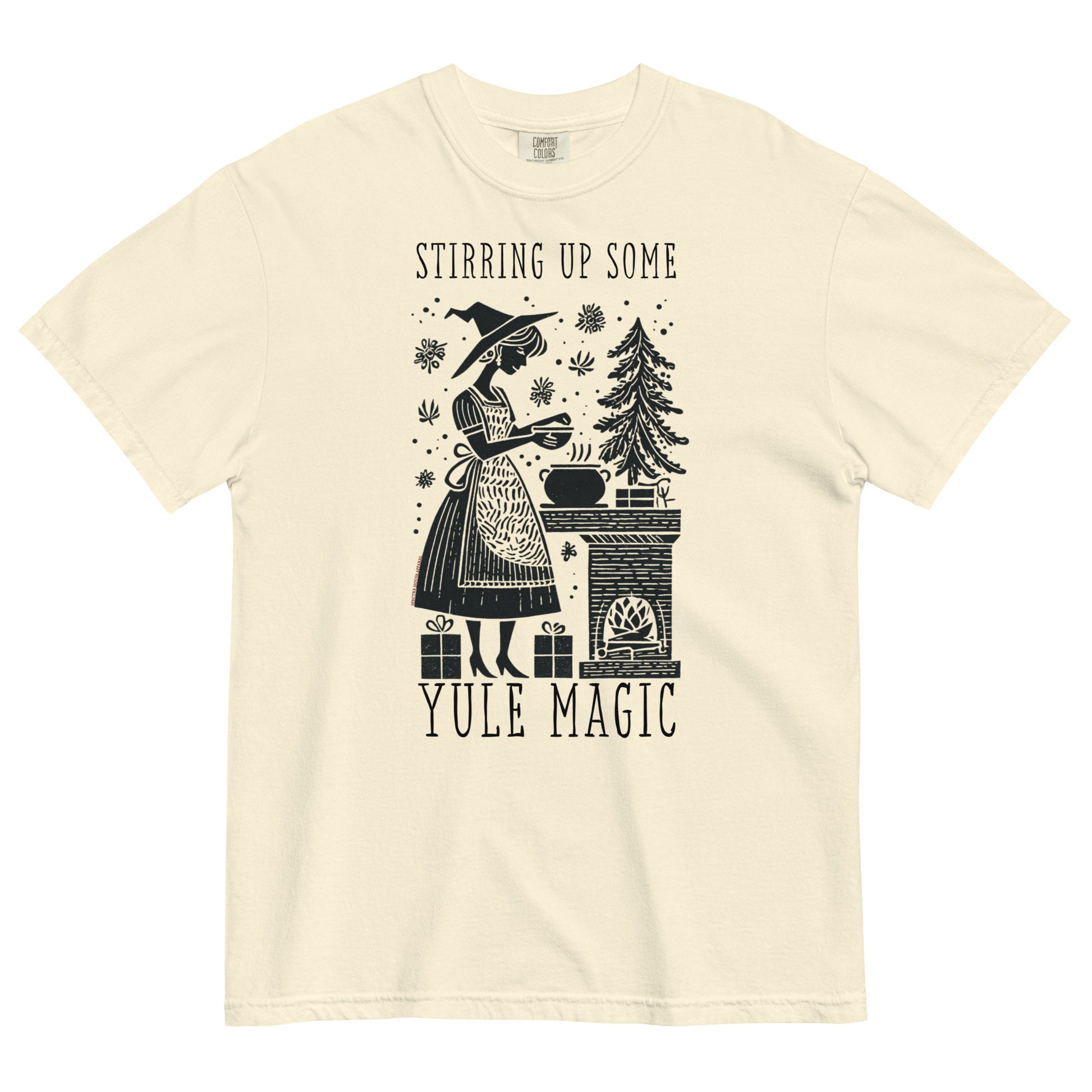 Cream t-shirt lay flat featuring vintage illustration of a witch stirring a cauldron beside a pine tree with text “Stirring Up Some Yule Magic,” a cozy witchy holiday tee.