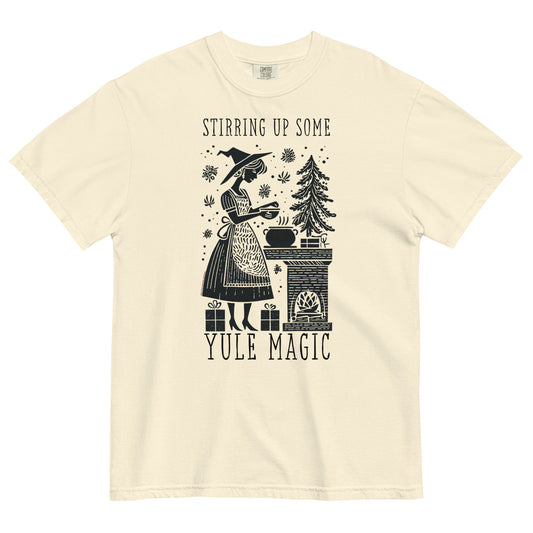 Cream t-shirt lay flat featuring vintage illustration of a witch stirring a cauldron beside a pine tree with text “Stirring Up Some Yule Magic,” a cozy witchy holiday tee.